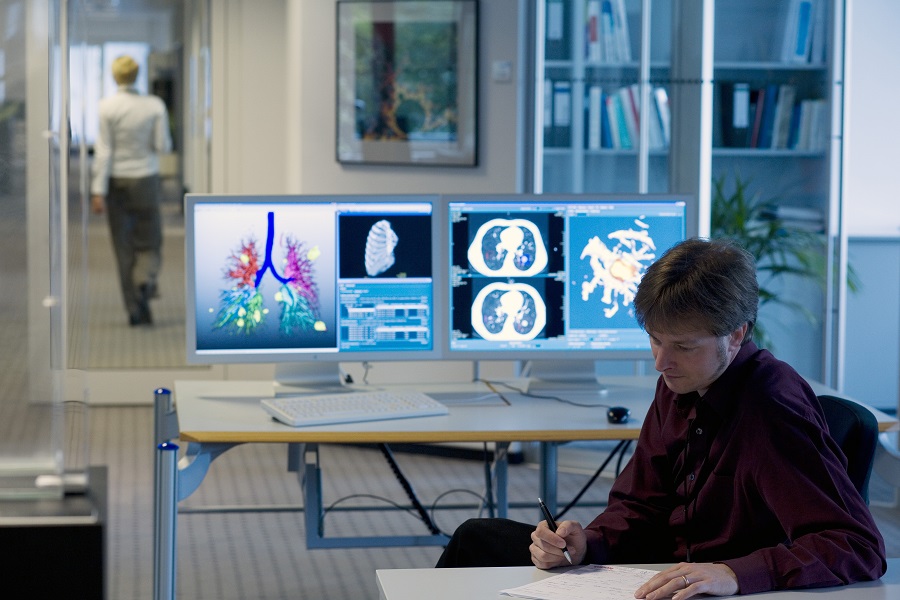 Ein Forscher macht sich Notizen, im Hintergrund sind zwei Monitore mit Ergebnissen der medizinischen Bildgebung.
