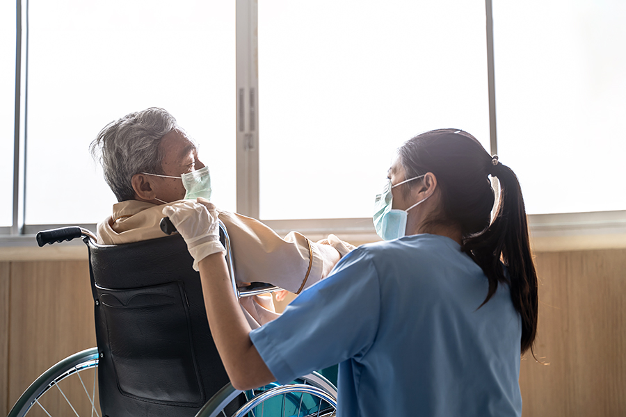 Patient im Rollstuhl mit asiatischer Pflegerin, beide tragen einen Mundschutz