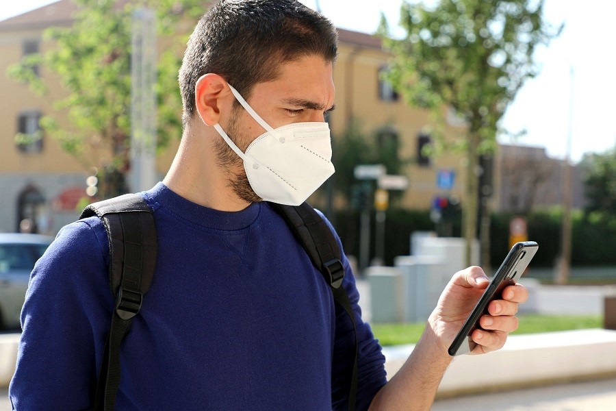 Junger Mann mit Mundschutz betrachtet Display eines Smartphones