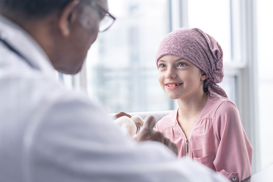 Arzt spricht im Krankenzimmer zu Kind mit rosafarbenem Kopftuch.