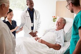 Ein Mann liegt in einem Krankhausbett und mehrere Personen mit unterschiedlicher Krankenhausbekleidung stehen um ihn herum