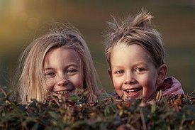 Zwei lachende Kinder mit vom Wind zerzausten Haaren schauen über eine Hecke