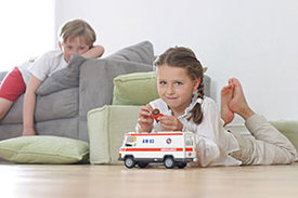 Ein Junge liegt auf dem Sofa und beobachtet ein Mädchen, das auf dem Boden spielt. 