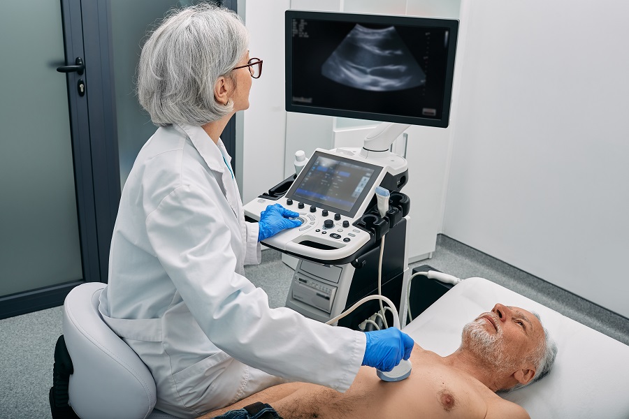 Eine Ärztin sitzt vor einem Ultraschallgerät und schallt die Brust eines Mannes, der neben ihr auf einer Liege liegt.