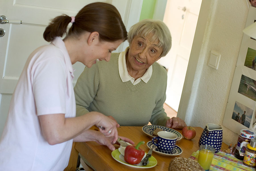 Eine Altenpflegerin unterstützt eine ältere Dame beim Frühstück im privaten Wohnumfeld.