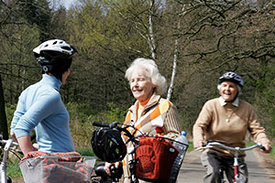 Eine ältere Dame fährt auf dem Fahrrad an zwei Personen vorbei, die sich unterhalten.